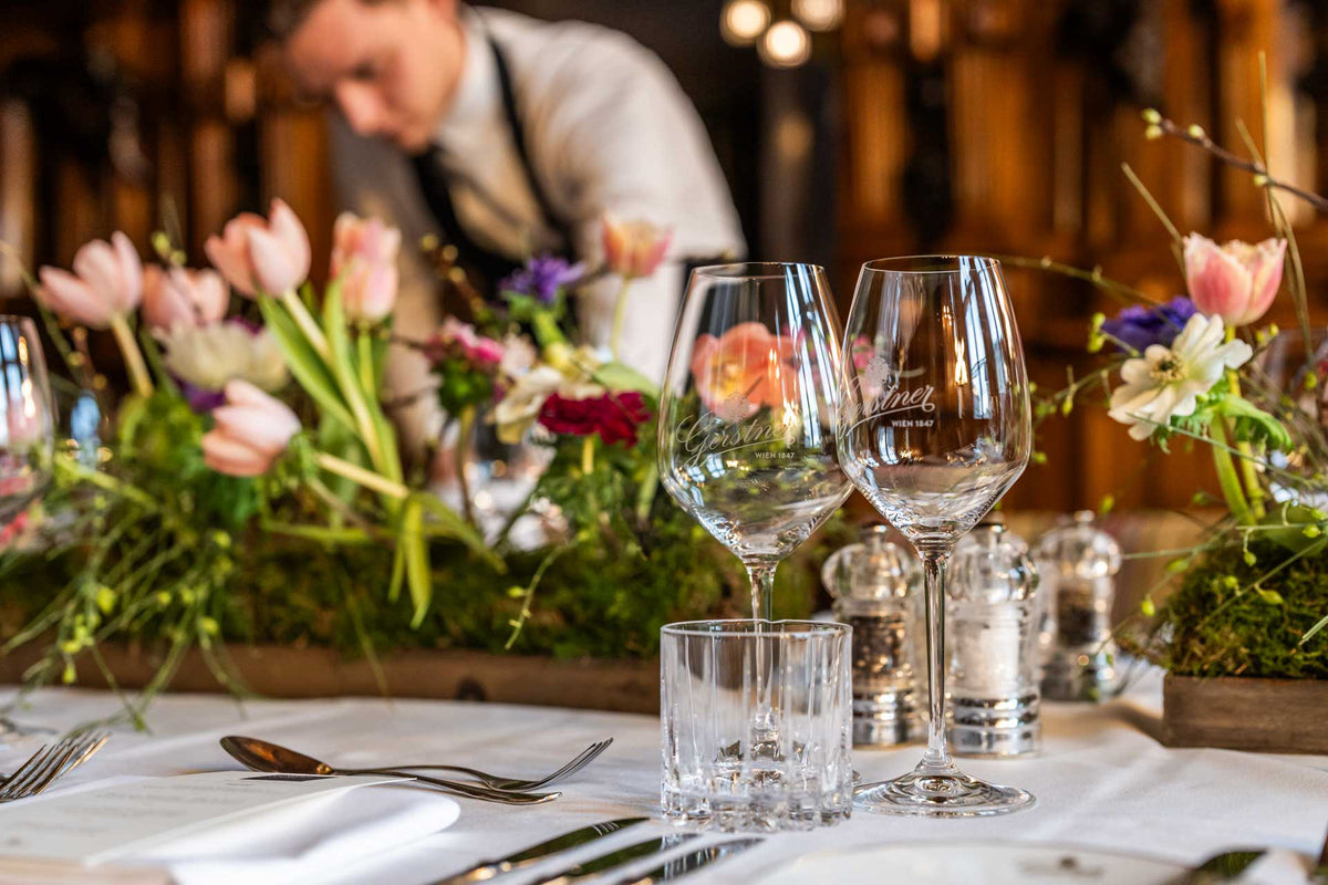 Tisch mit Besteck, zwei Weingläsern, Wasserglas, Salz- und Pfefferstreuern sowie mehreren Blumenarrangements und Kellner im Hintergrund