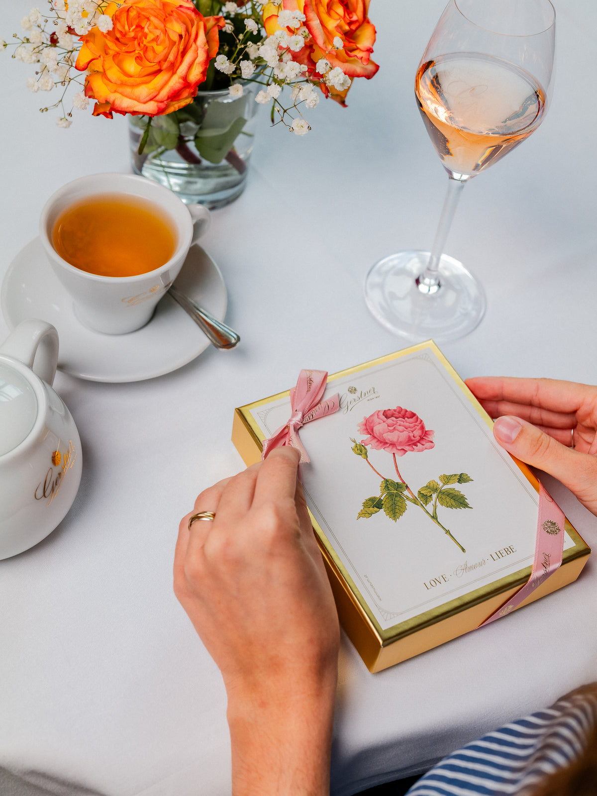 Beide Hände einer Person öffnen eine elegante Schachtel mit rosa Schleife neben Tee, einem Glas Wein und Blumen auf einem gedeckten Tisch